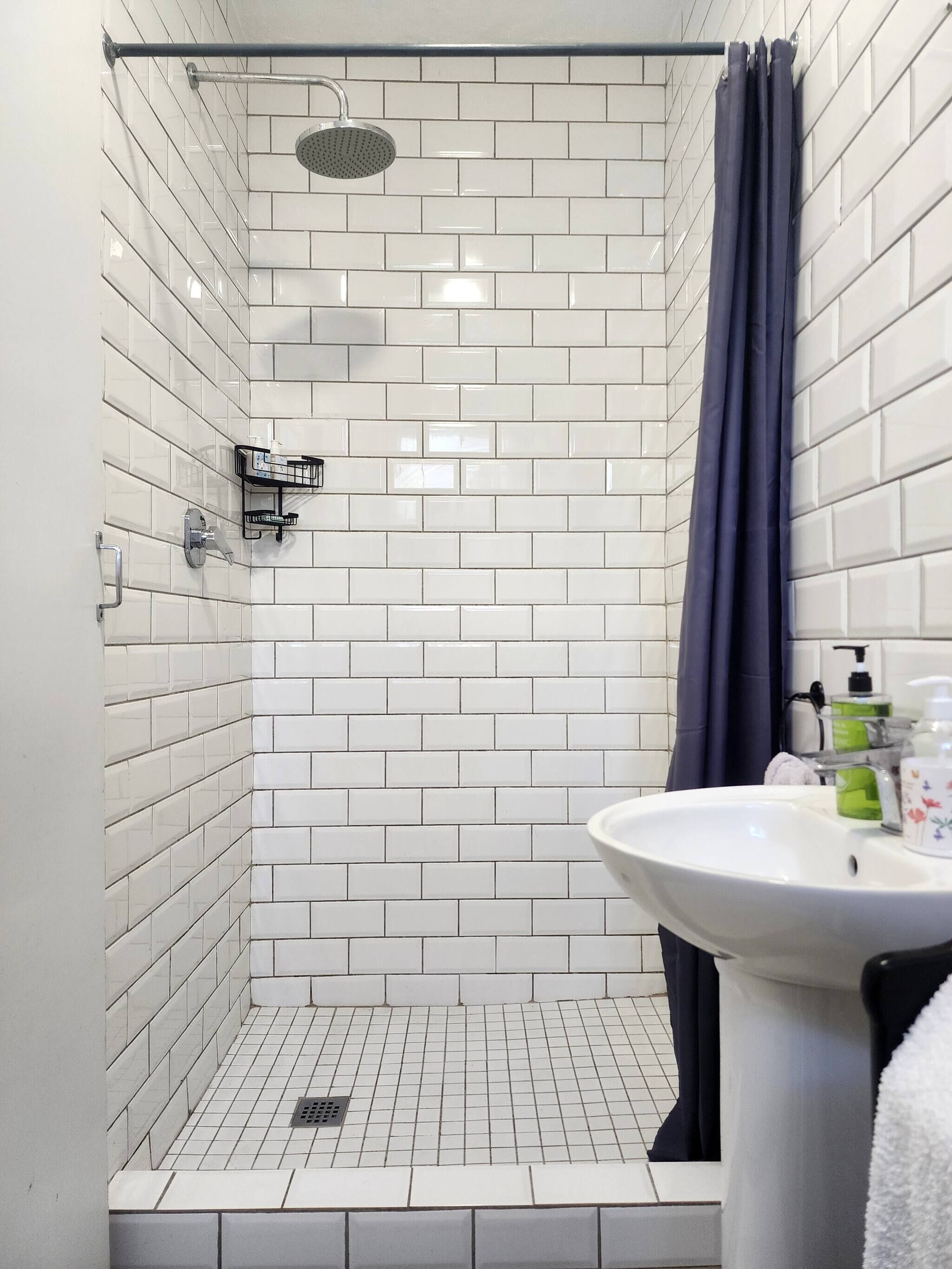 Une douche aux carreaux blancs, un pommeau de douche, un rideau foncé et un lavabo avec des articles de toilette.