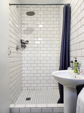 A shower with white tiles, a showerhead, a dark curtain, and a sink with toiletries.