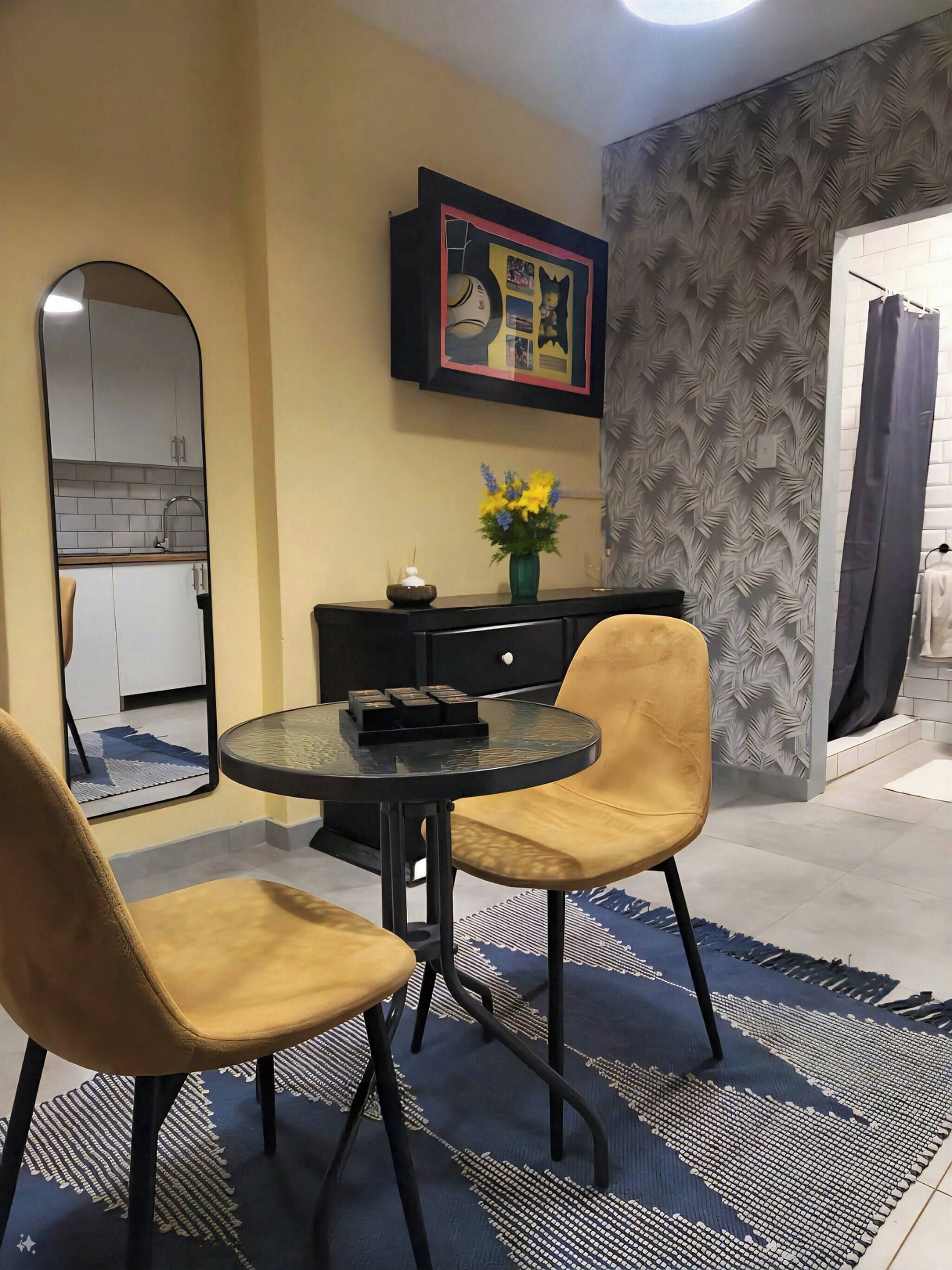 Un coin repas avec une table ronde, deux chaises, un vase de fleurs, un tableau encadré et une cuisine visible à travers une arche.