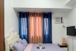 A bedroom with a bed, curtains, an air conditioning unit, a television, and a bedside table. - Grand Riviera Suites – Manila Bay Stay (Manila)
