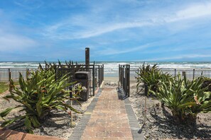 Ein Weg, der zu einem Strand mit Zaun, Pflanzen und Mülleimer führt.