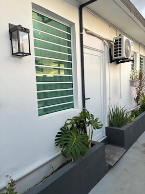 Fachada blanca con una puerta, una ventana con persianas horizontales, una lámpara de pared y una jardinera con plantas.