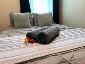 Una cama con un edredón estampado, dos toallas enrolladas y una sola hoja de otoño.