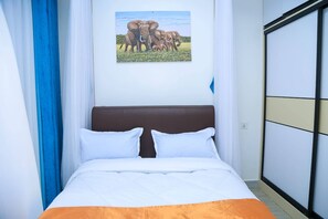 A bed with white and orange bedding, a brown headboard, and a painting of elephants on the wall. - 1 room in a 2 bedroom apt near the airport and SGR train station Nairobi Kenya (Nairobi)