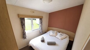 A bedroom with a bed, a window with curtains, and a wooden headboard. - MKRJ COMFORT– Parkdean Camber Sands – Sleeps 8 - Pet Friendly - lbl5 (Camber)