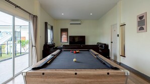 Una mesa de billar con bolas, un televisor y un sofá en una habitación con un ventanal grande y un equipo de aire acondicionado.