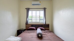 Una cama individual con cabecera, una ventana con cortinas, un aire acondicionado de pared y una mesita de noche pequeña con lámpara.