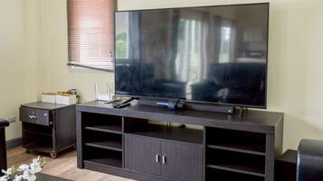 A modern living room with a flat-screen TV on a dark wooden entertainment unit, a small side table with a vase of flowers, and a window with blinds.