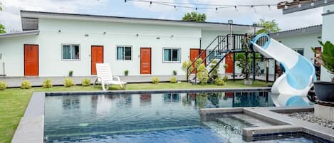 A swimming pool with a slide, a white building with red doors, and a staircase.