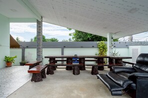 An outdoor patio with a long wooden table and benches, surrounded by potted plants and a black leather sofa.