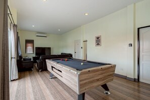 A pool table in a room with a sofa, a television, and an air conditioning unit.