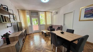 Ein Zimmer mit Holzdielen in Fischgrätmuster, einem Esstisch mit Stühlen, einer Couch, einem Fernseher und einem Bücherregal.