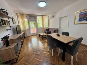 Ein Zimmer mit Holzdielen in Fischgrätmuster, einem Esstisch mit Stühlen, einer Couch, einem Fernseher und einem Bücherregal.