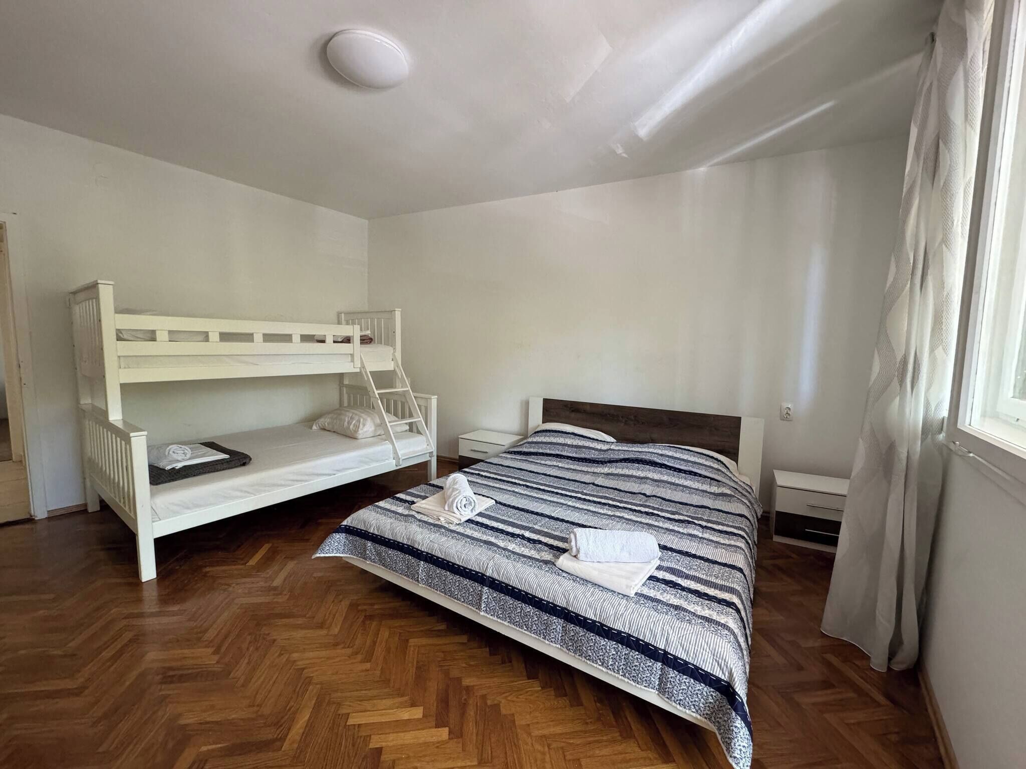 A room with a bunk bed, a single bed, a wooden floor, a window with curtains, and a ceiling light.