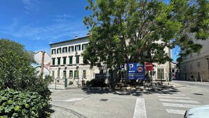Straßenansicht mit einem weißen Auto am Straßenrand, einem Gebäude mit einem Schild und einem Baum im Vordergrund.