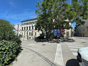 Straßenansicht mit einem weißen Auto am Straßenrand, einem Gebäude mit einem Schild und einem Baum im Vordergrund.