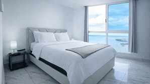 Un dormitorio moderno con una cama grande, una mesita de noche, una lámpara y vista al mar a través de un ventanal amplio.