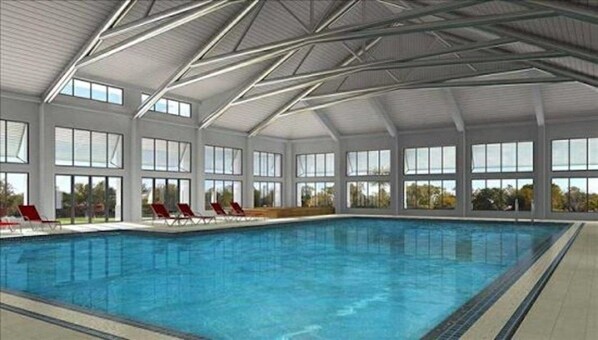 An indoor swimming pool with clear glass walls, red lounge chairs, and a high ceiling with metal beams.