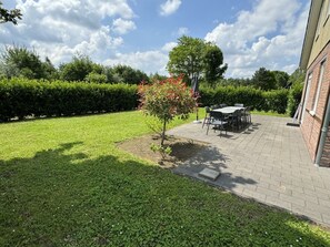 Een terras met een tafel en stoelen, een kleine boom en een haag in een verzorgde tuin.