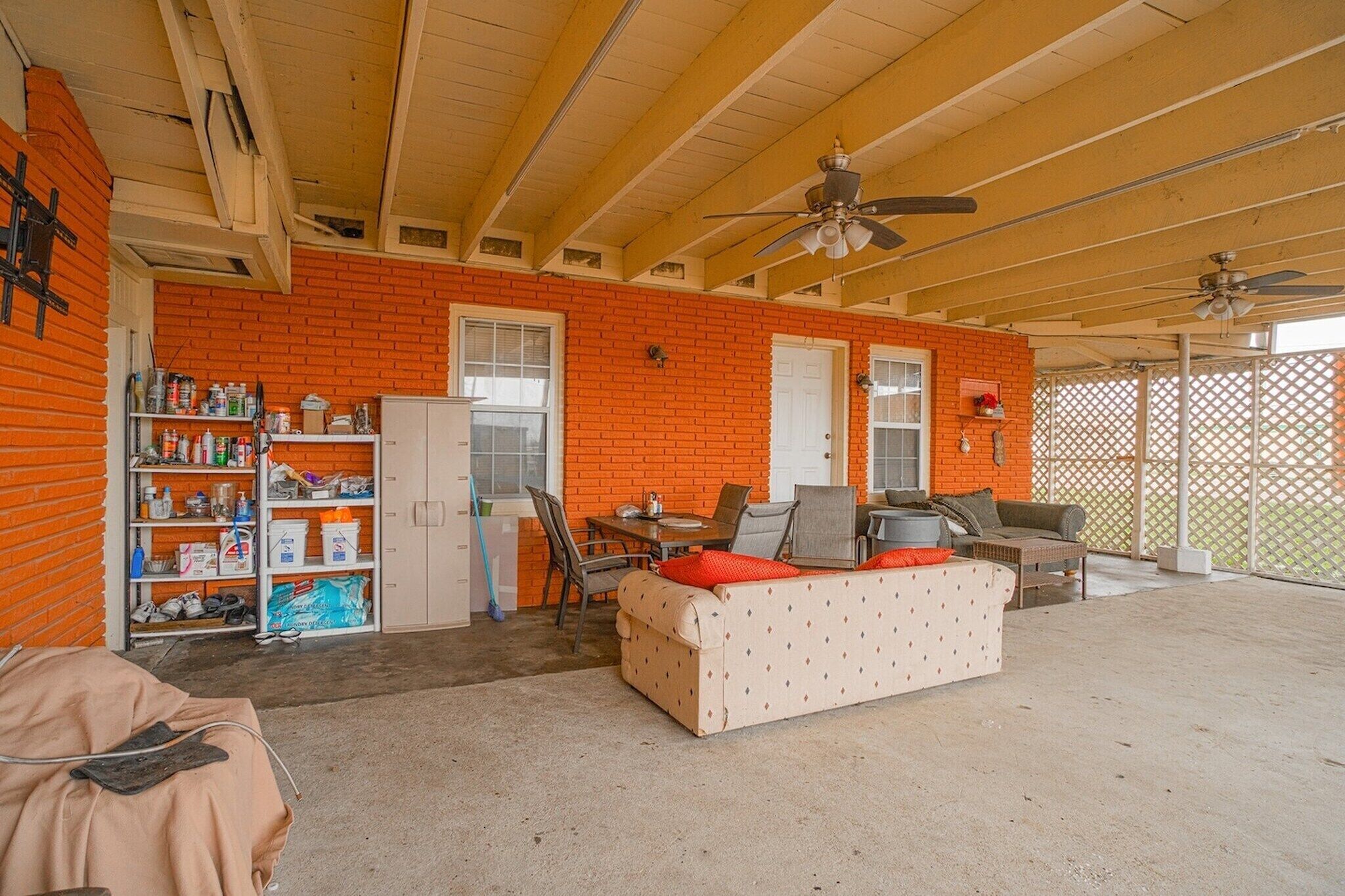 Ein überdachter Patio-Bereich mit Backsteinmauer, einem weißen Sofa, einem Esstisch und Deckenventilatoren.
