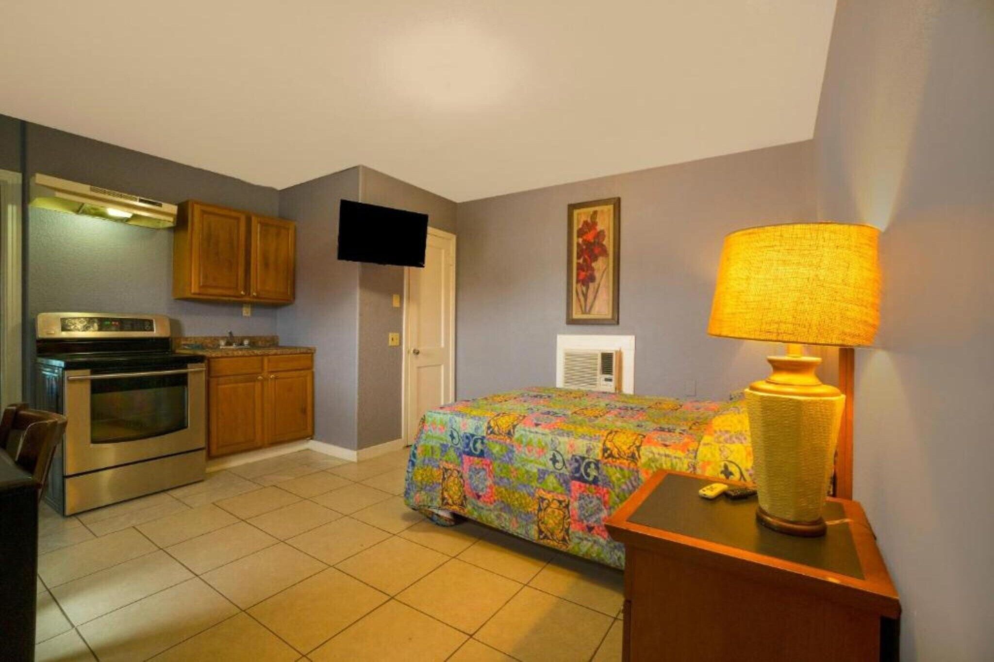 A compact hotel room with a kitchenette, a bed with a colorful bedspread, a television, and a lamp.