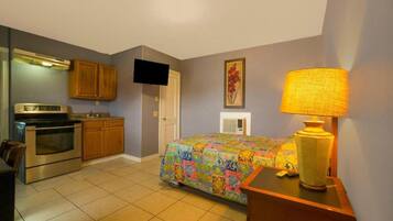 A compact hotel room with a kitchenette, a bed with a colorful bedspread, a television, and a lamp.