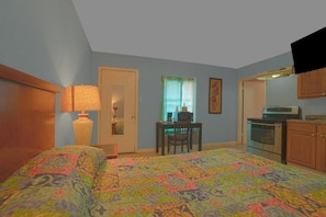 A bedroom with a bed, a desk, a chair, a television, and a kitchenette.