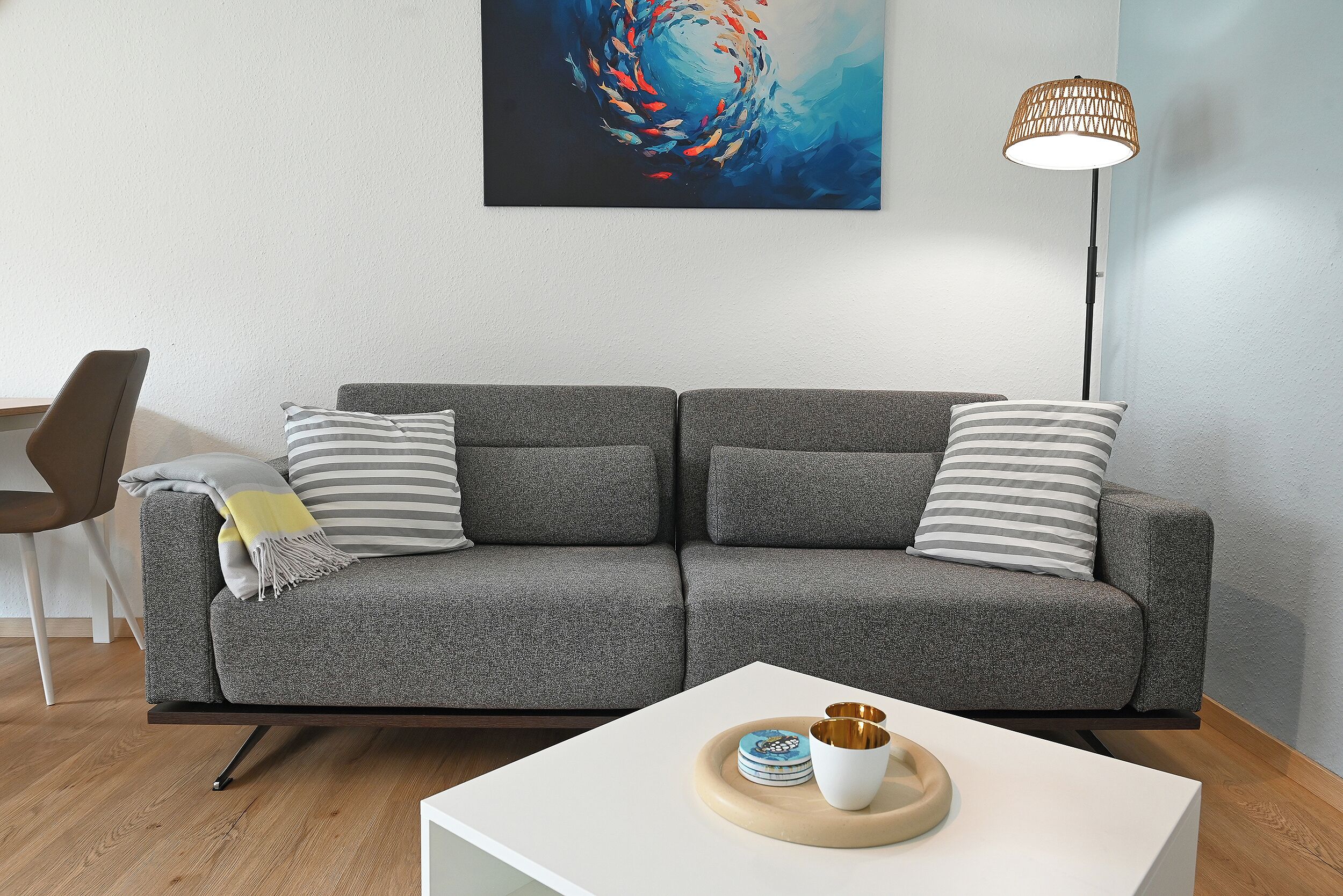 Een moderne woonkamer met een grijze bank, gestreepte kussens, een bijzettafeltje met decoratieve objecten, een vloerlamp en een kunstwerk aan de muur.