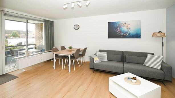 Ein modernes Wohnzimmer mit Essbereich, einem grauen Sofa, einem weißen Couchtisch und einem großen Fenster mit Blick auf Gebäude.