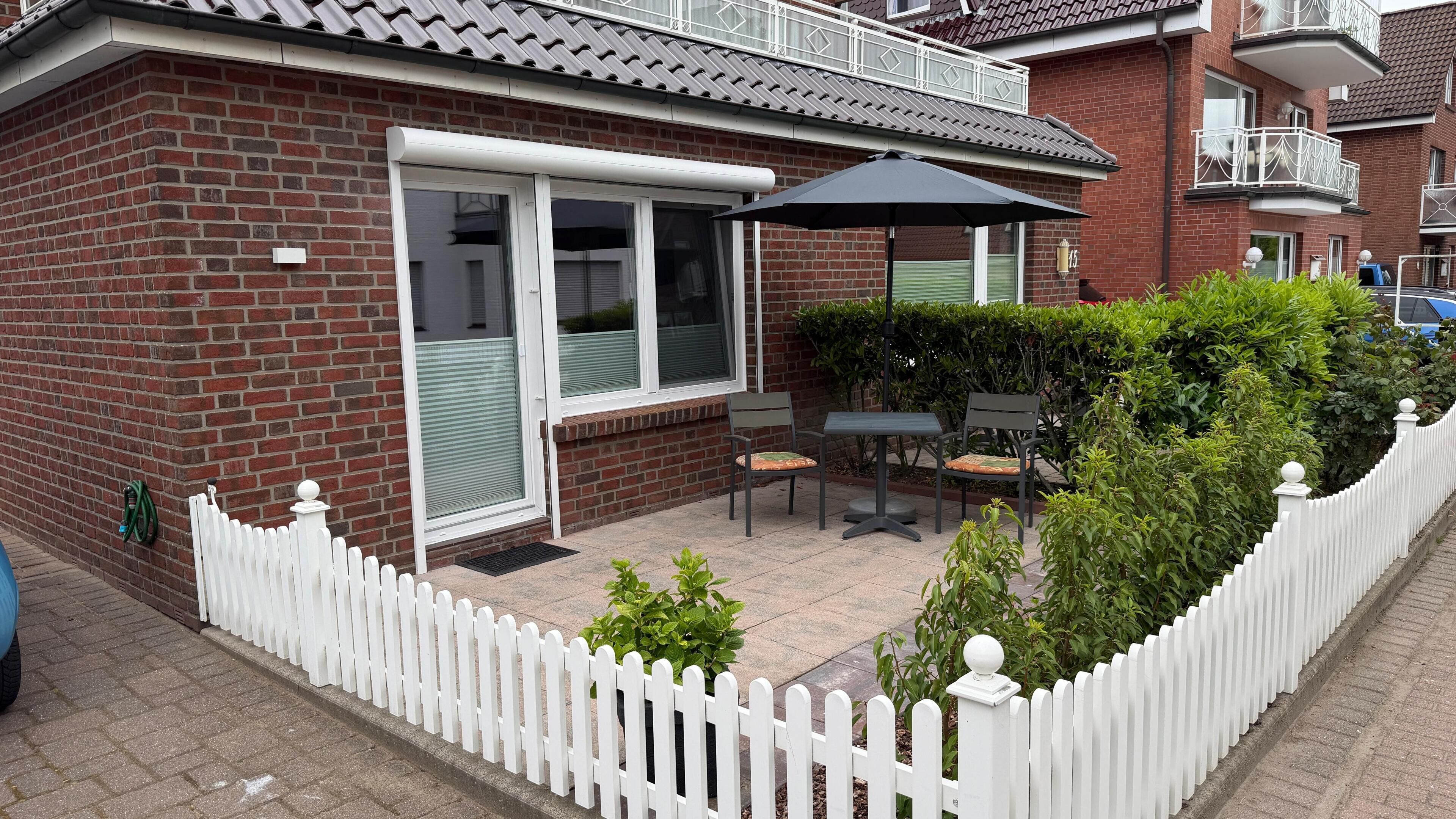 A residential building with a brick facade, a white picket fence, and a patio area with outdoor furniture and a table.