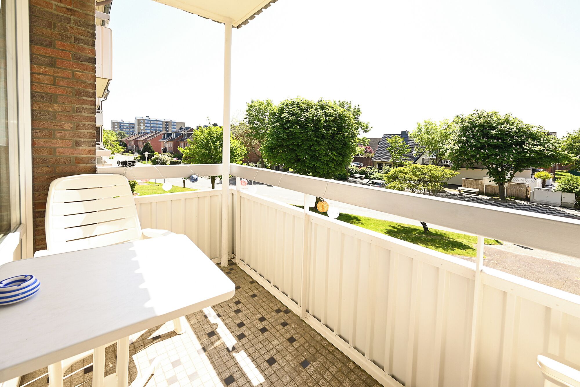 Un balcón con una mesa y sillas blancas, vista a una zona residencial con casas y vegetación.