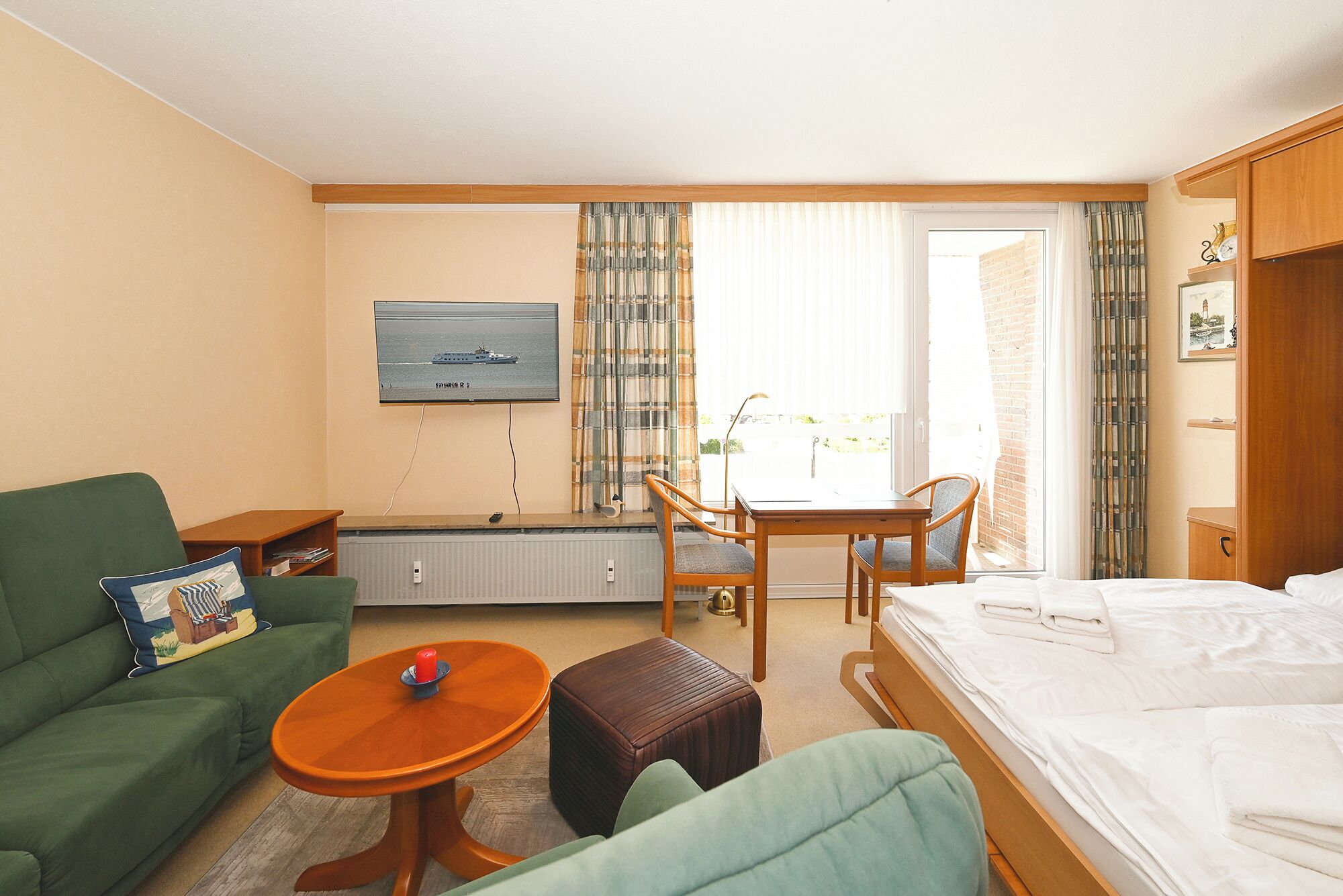 Habitación de hotel con cama, sofá verde, mesa de madera, televisor y ventana con cortinas.