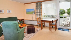 A living room with a green sofa, a wooden table with chairs, a television, and a view of a balcony with outdoor furniture. - Glück vacation apartment with balcony and lots of sunshine (Büsum)