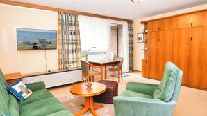 A living room with a green sofa, a green armchair, a wooden dining table, and a view of the sea through a window. - Glück vacation apartment with balcony and lots of sunshine (Büsum)