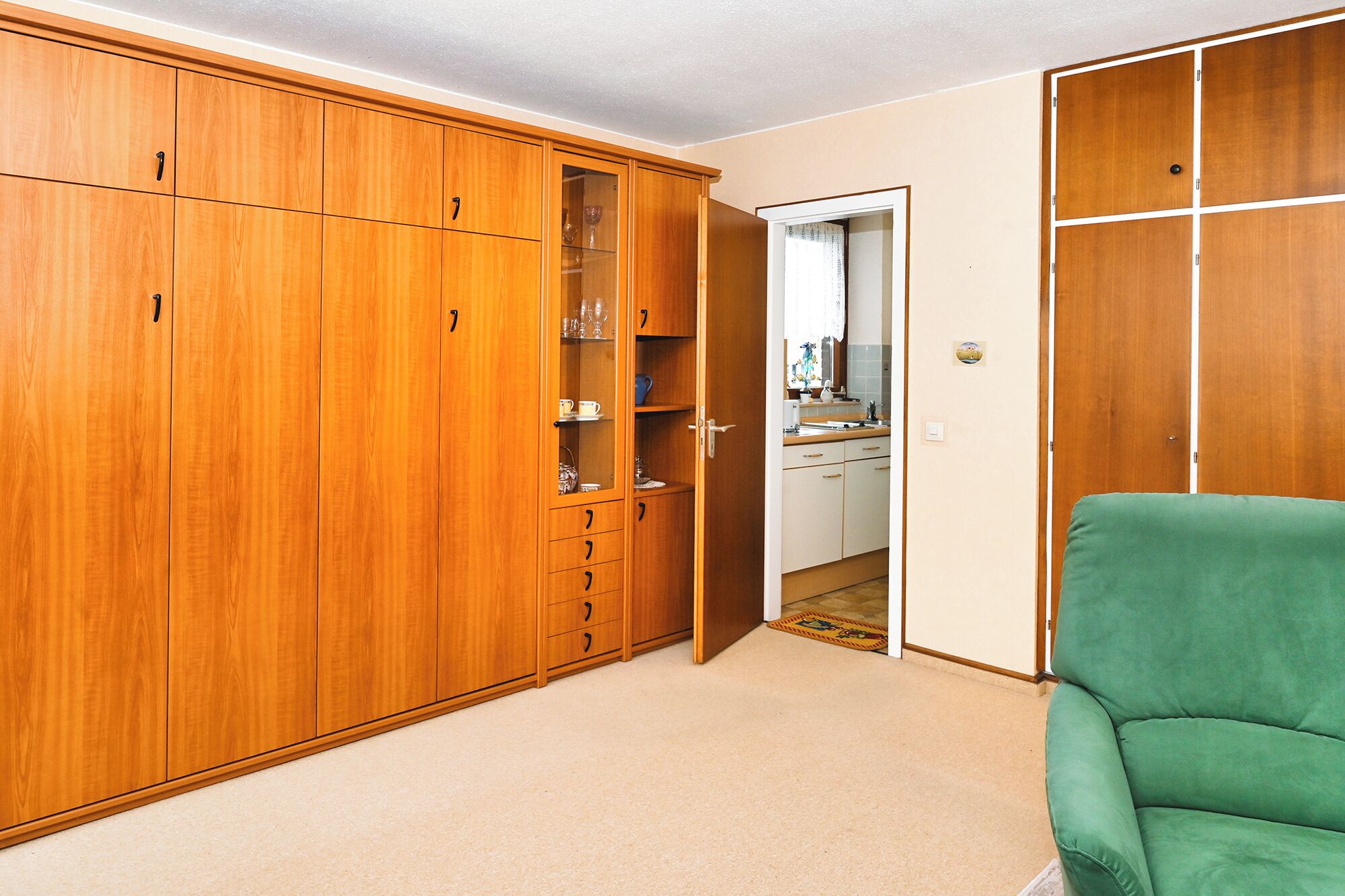 A room with a large wooden wardrobe, a green armchair, and a kitchen area visible through an open door.