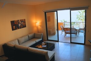Un salón con sofá, una mesa de centro, una lámpara y un ventanal corredero que da a un balcón con mobiliario exterior.