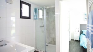 Une salle de bain moderne avec une douche à parois de verre, une baignoire blanche et une porte visible donnant sur une chambre à coucher équipée d’un lit et d’une table de chevet.