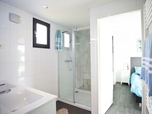A modern bathroom with a glass-enclosed shower, a white bathtub, and a visible door leading to a bedroom with a bed and a nightstand. - Cube Exclusive 6 (Nijkerk)