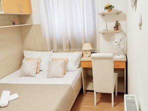 Ein Schlafzimmer mit einem Bett, einem Schreibtisch, einem Sessel, einer Lampe, einem Ventilator und einem Regal mit Dekoration.