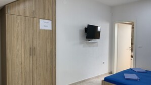 Quarto com guarda-roupa de madeira, televisão na parede, cama com colcha azul e mesa branca.