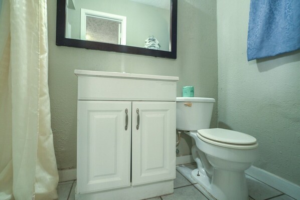 Un baño con inodoro blanco, un mueble blanco con dos puertas, un espejo sobre el mueble y una toalla azul colgada en la pared.