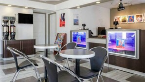 Moderni hotellin aula, jossa on pyöreitä pöytiä, mustia tuoleja, kahvinkeitin ja televisio, jolta näkyy uutiskanava.
