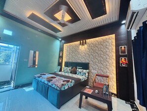 A modern bedroom with a large bed, a bedside table, a chair, a window, and a wall-mounted air conditioning unit. - Vishnu Priya Guest House Ayodhya (Ayodhya)