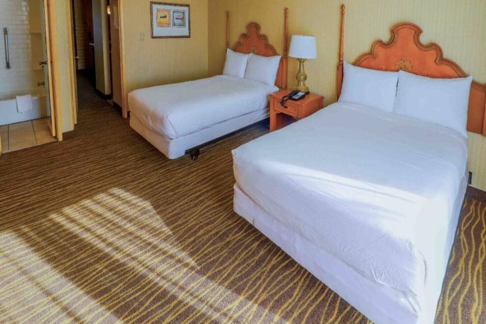 Camera d'albergo con due letti, una testiera in legno, un comodino con una lampada e un pavimento rivestito di moquette.