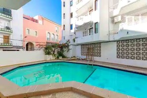 Una piscina con suelo de baldosas, rodeada de edificios con balcones y una valla de celosía.