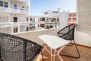 Un balcón con dos sillas de mimbre, una mesa de metal blanco y vistas a edificios cercanos.