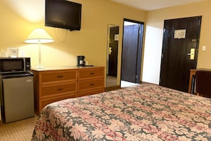 Habitación de hotel con cama, escritorio con lámpara, microondas, cafetera y televisión en la pared.