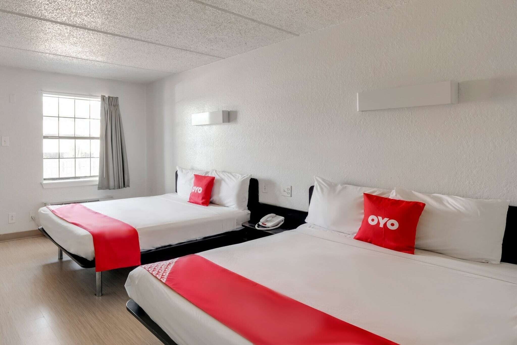 Two beds with white bedding and red accents, a window with curtains, a wall-mounted light, and a phone on the nightstand.