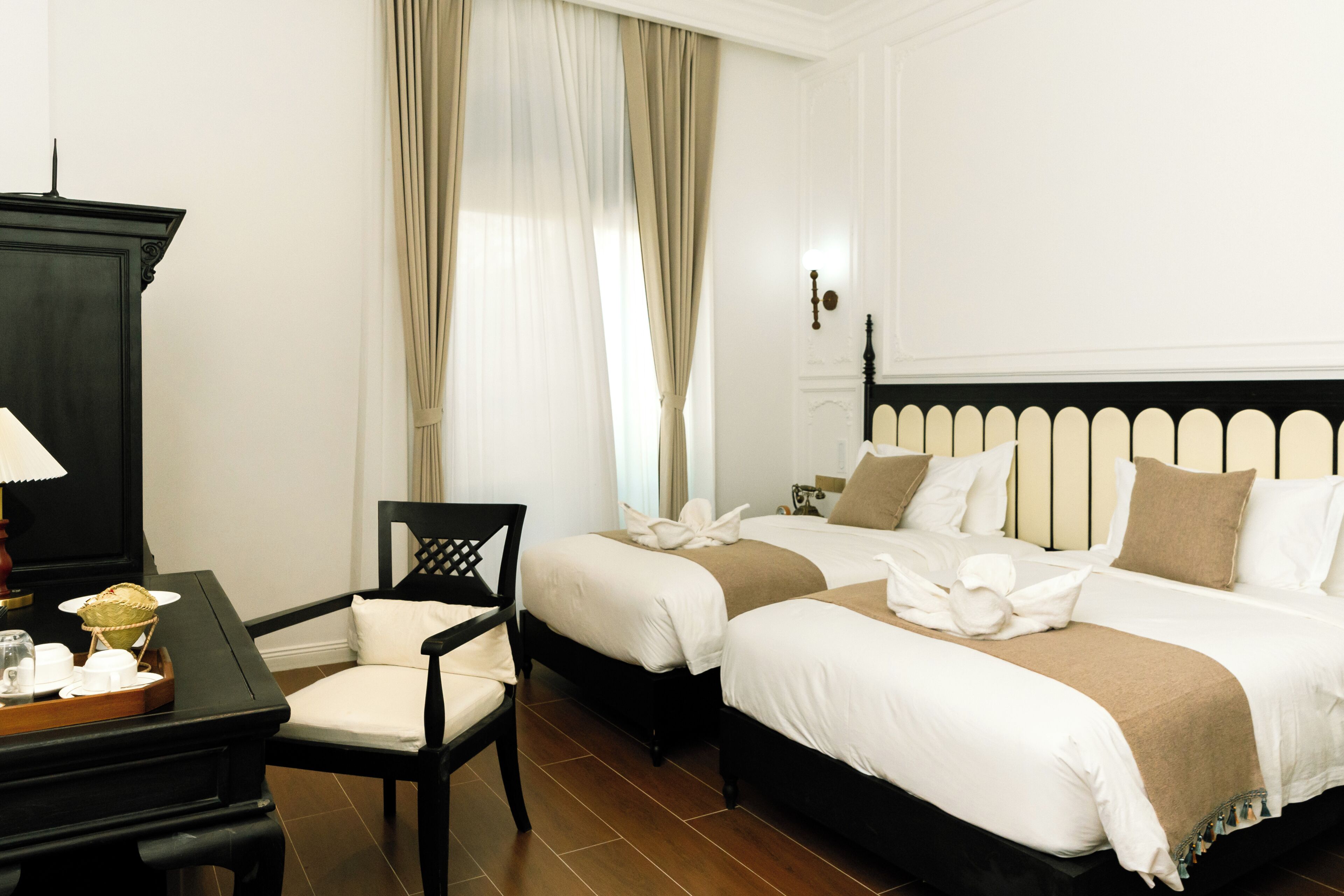 Habitación de hotel con dos camas, un escritorio con lámpara, una silla y un ventanal con cortinas.