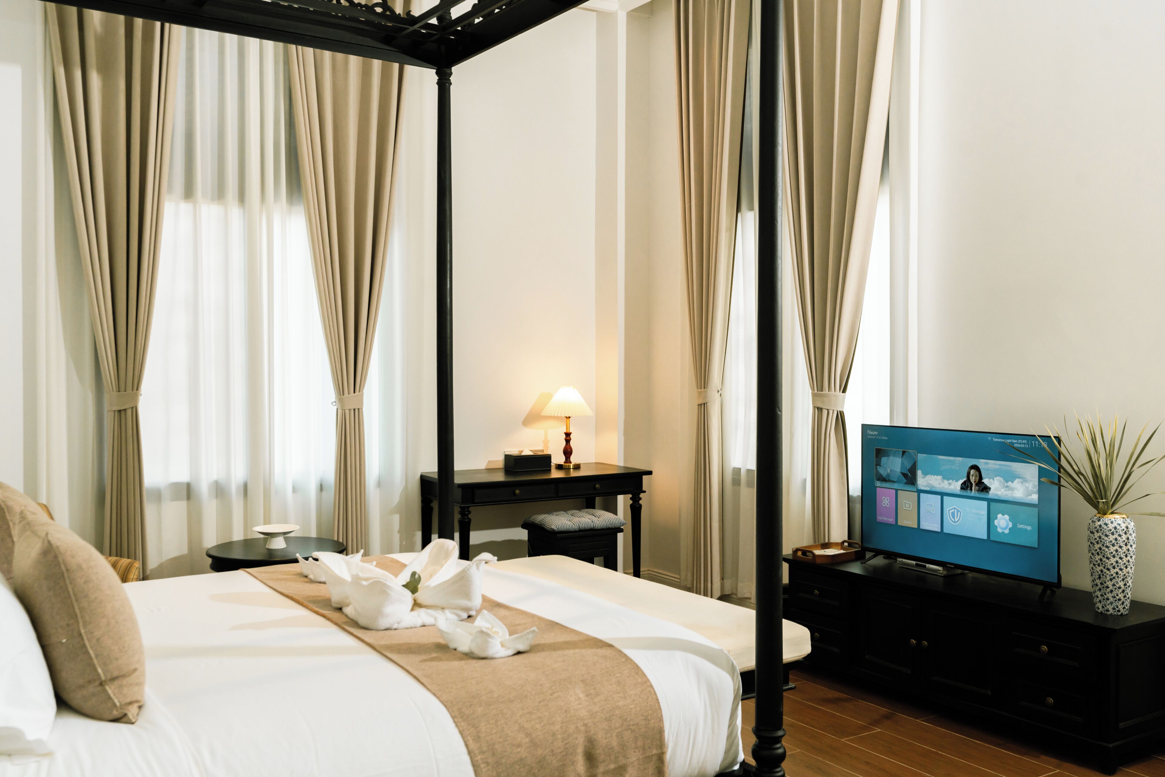A hotel room with a four-poster bed, a flat-screen TV, a desk with a lamp, and a potted plant.